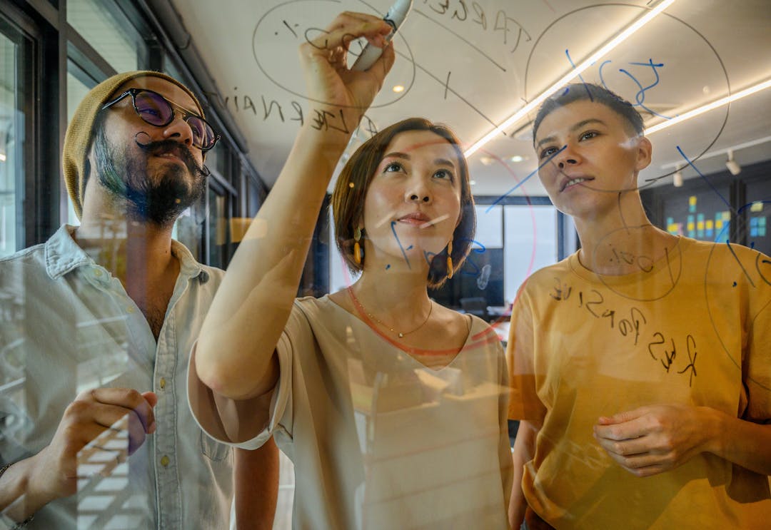 En kvinnlig och två manliga kollegor brainstormar på kontoret och samarbetar på en glasskiva med markörer, vilket främjar teamwork och kreativitet.