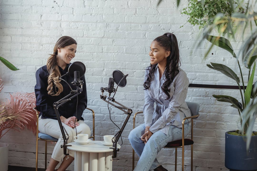 Två leende kvinnor spelar in en podcast tillsammans i ett rum med vita tegelväggar och växter.