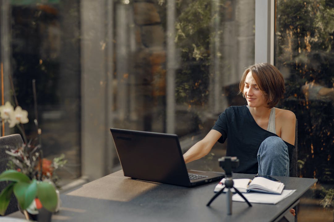 Kvinna med innehåll arbetar med bärbar dator vid ett soligt skrivbord med en smartphone på ett stativ och anteckningsböcker i närheten.