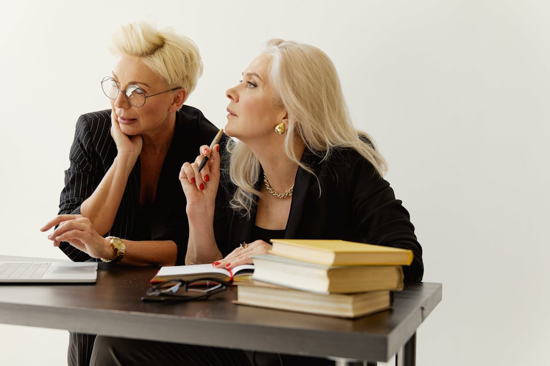 Två affärskvinnor i diskussion, med böcker och en bärbar dator på skrivbordet, mot en vit bakgrund.